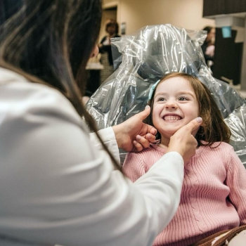 A dentist examines a smiling young girl sitting in a dental chair covered with plastic.