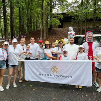 A group of people in white shirts stands together outdoors, holding a “Brightworks” banner, with some dressed in costumes and trees visible in the background.