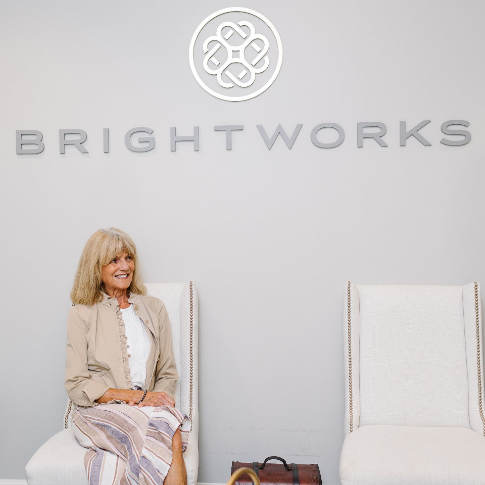 A woman sits on a white chair next to an empty chair beneath a wall sign that reads "BRIGHTWORKS.