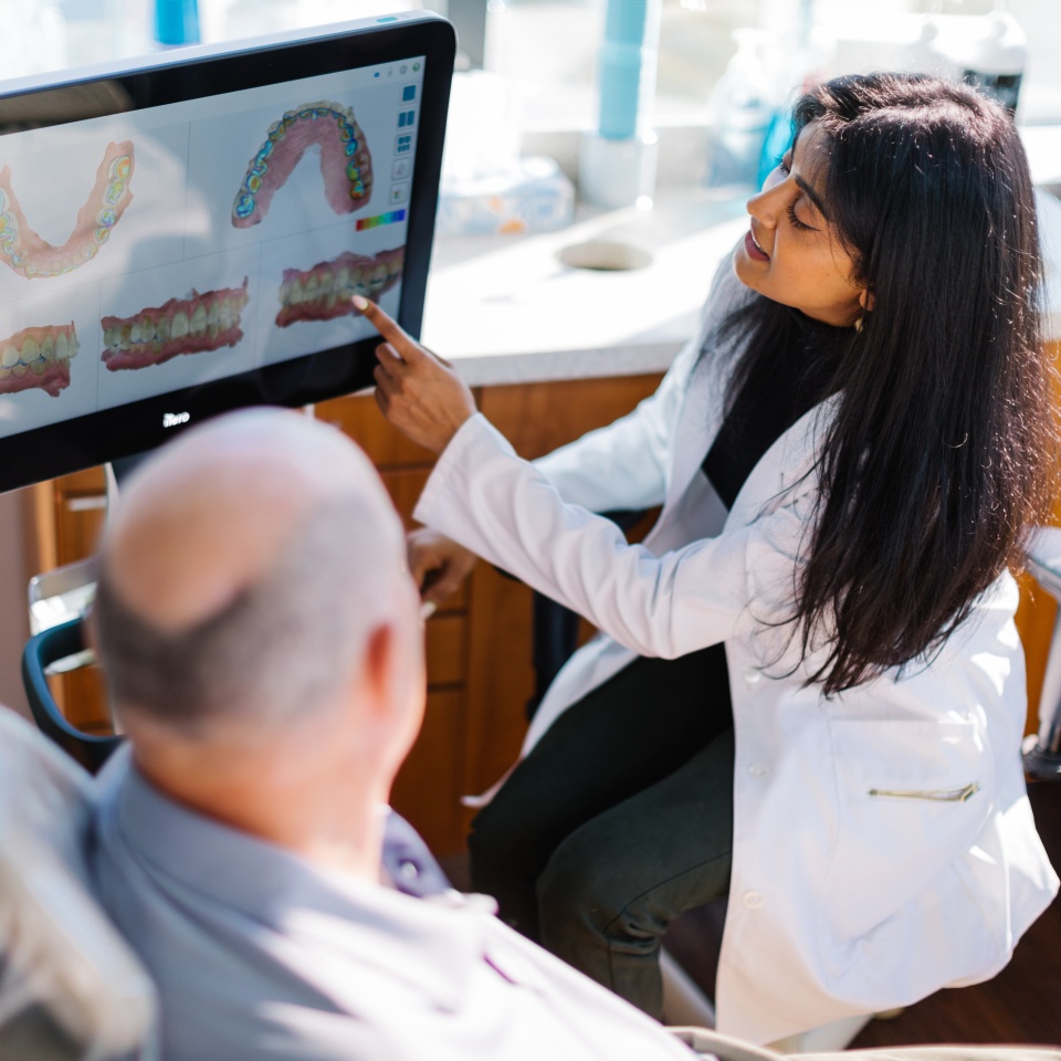 A dentist in a white coat shows a dental scan on a computer monitor to a seated patient in a dental office.