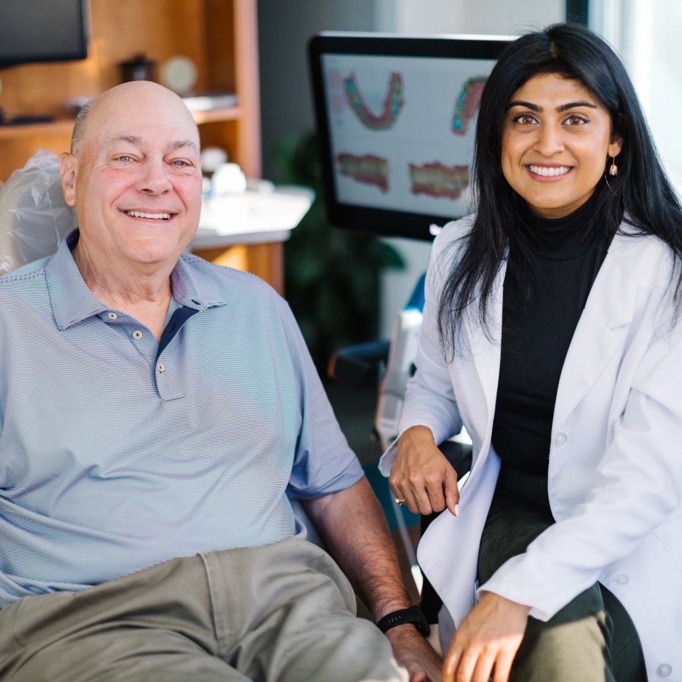An older man sits in a dental chair smiling next to a female dentist in a white coat, with dental images displayed on a monitor in the background.