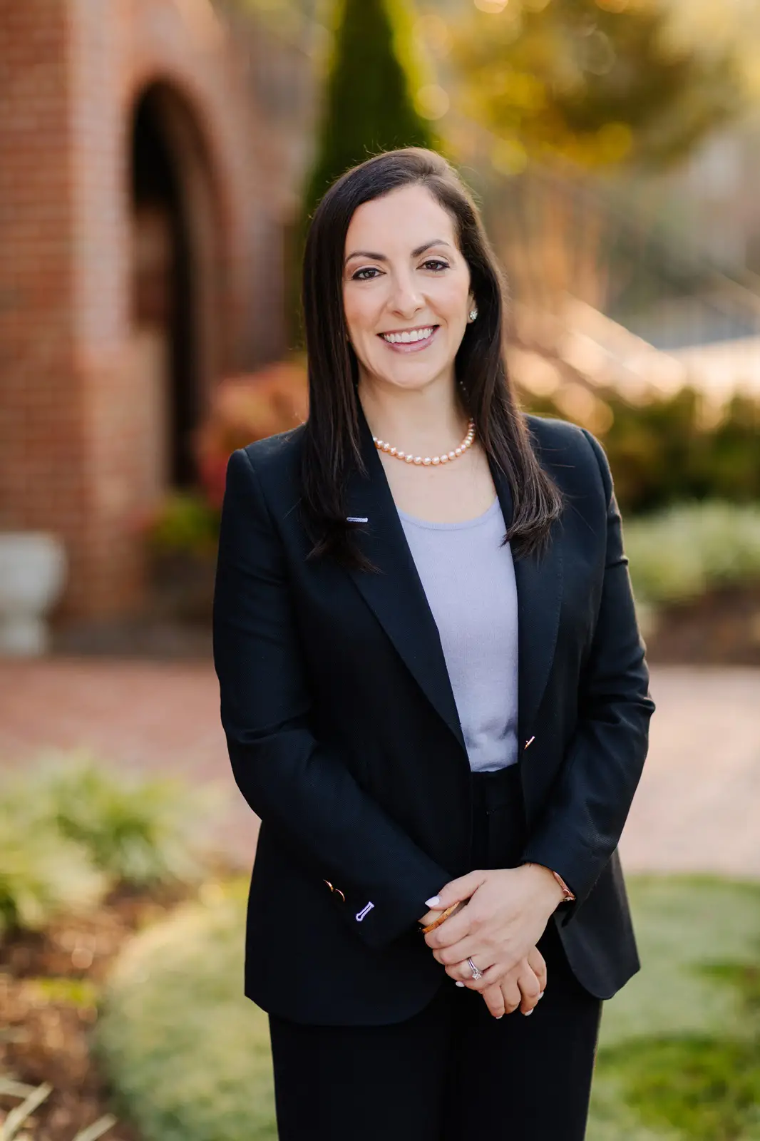 A woman in a dark suit and white blouse stands outside, smiling at the camera, with a brick building and greenery in the background.