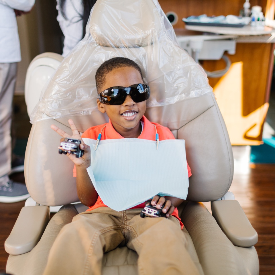 A young boy wearing sunglasses and a dental bib sits in a dentist's chair, smiling and holding toy cars while making a peace sign with his fingers.