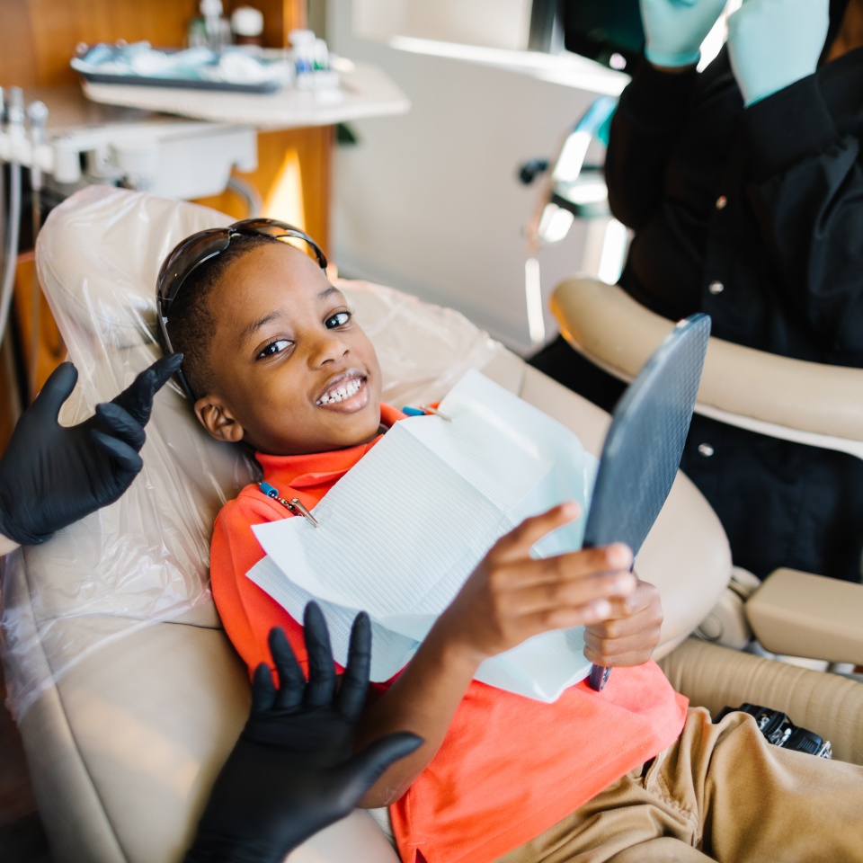 A boy sits in a dental chair, wearing a bib and sunglasses, smiling and holding a mirror, while dental professionals in gloves attend to him.