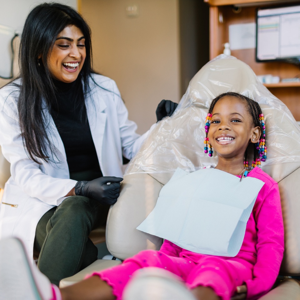 A dentist in a white coat sits beside a smiling young girl with beaded hair, seated in a dental chair and wearing a bib during a dental appointment.