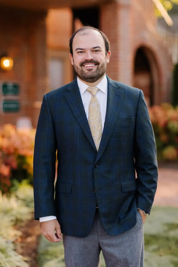 A man in a plaid suit jacket, white shirt, and patterned tie stands outdoors in front of a brick building and landscaped foliage, smiling at the camera.