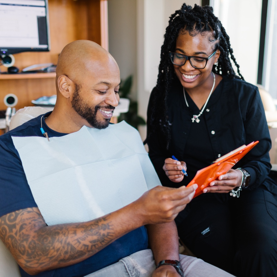 A man with a dental bib smiles while reviewing a clipboard with a female dental professional in an office setting.