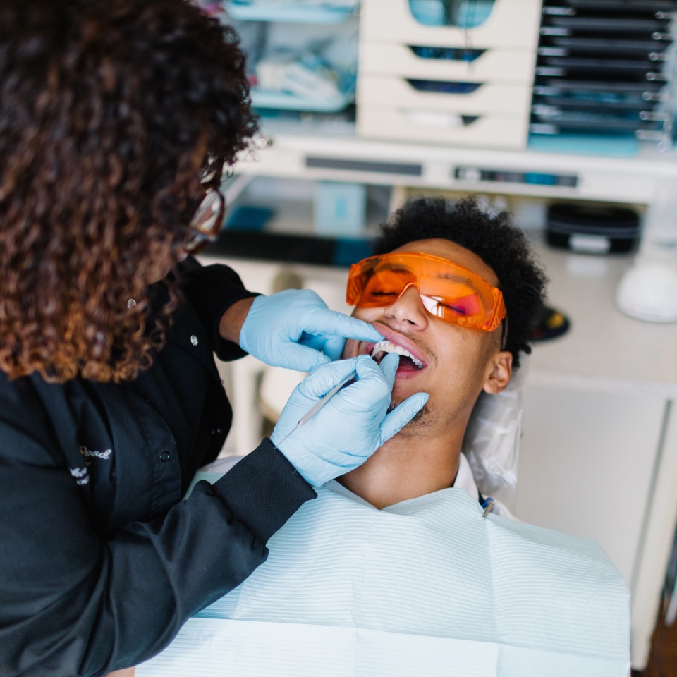 A dental professional wearing gloves examines a patient's teeth as the patient reclines in a dental chair and wears protective orange glasses.