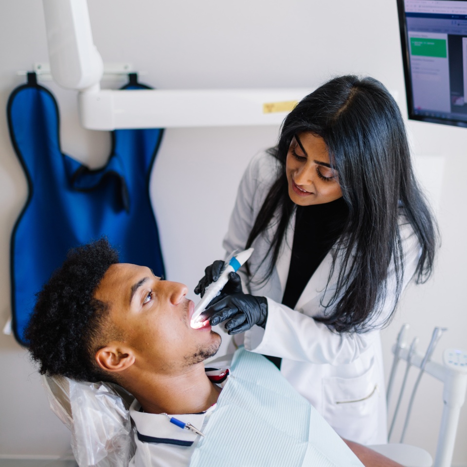 A dentist examines a patient's teeth using a dental tool while the patient sits in a dental chair. A monitor and dental apron are visible in the background.