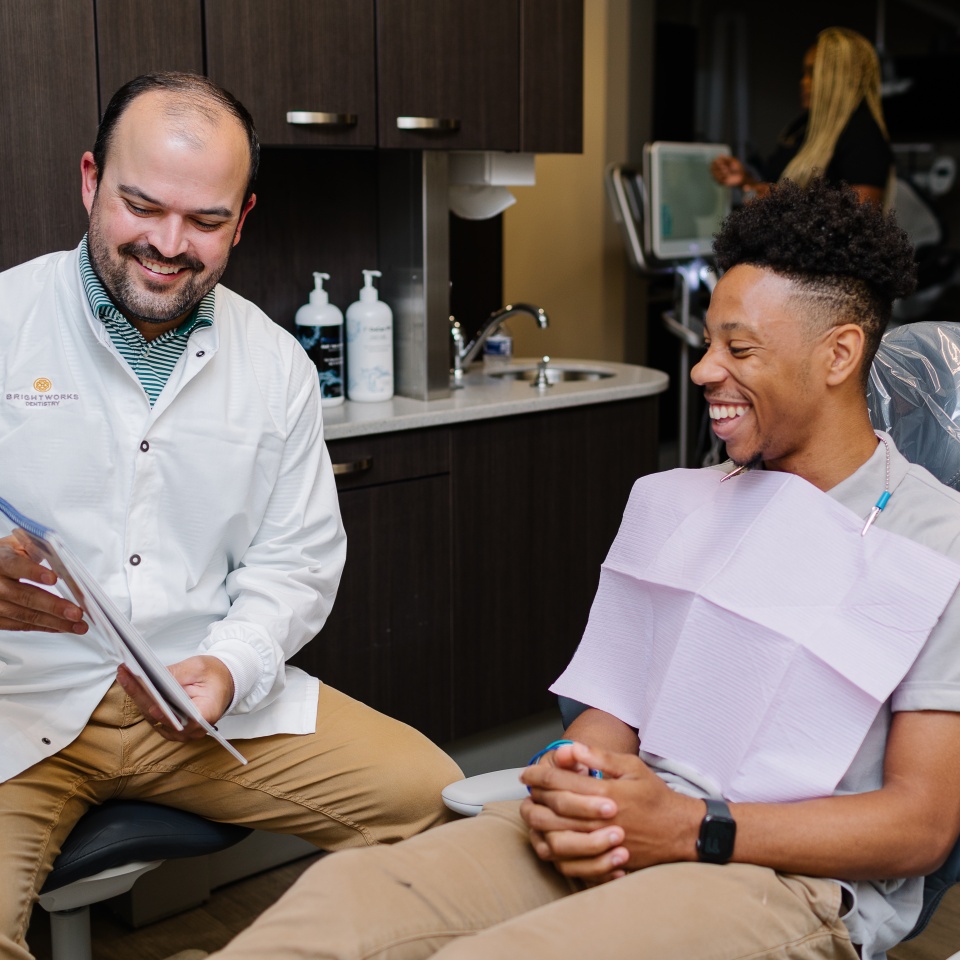 A dentist shows a patient a document while both smile in a dental office; the patient is seated wearing a dental bib.