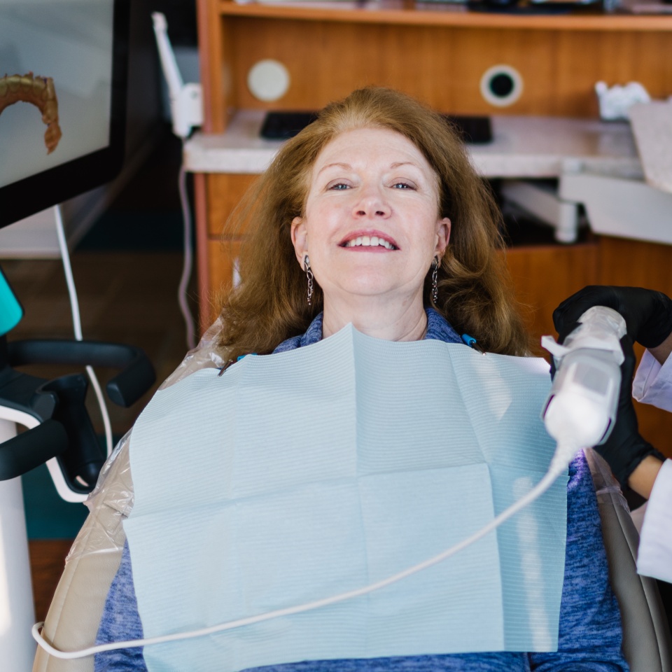A woman with red hair sits in a dental chair, wearing a blue bib, while a gloved dental professional uses a handheld device in her mouth.