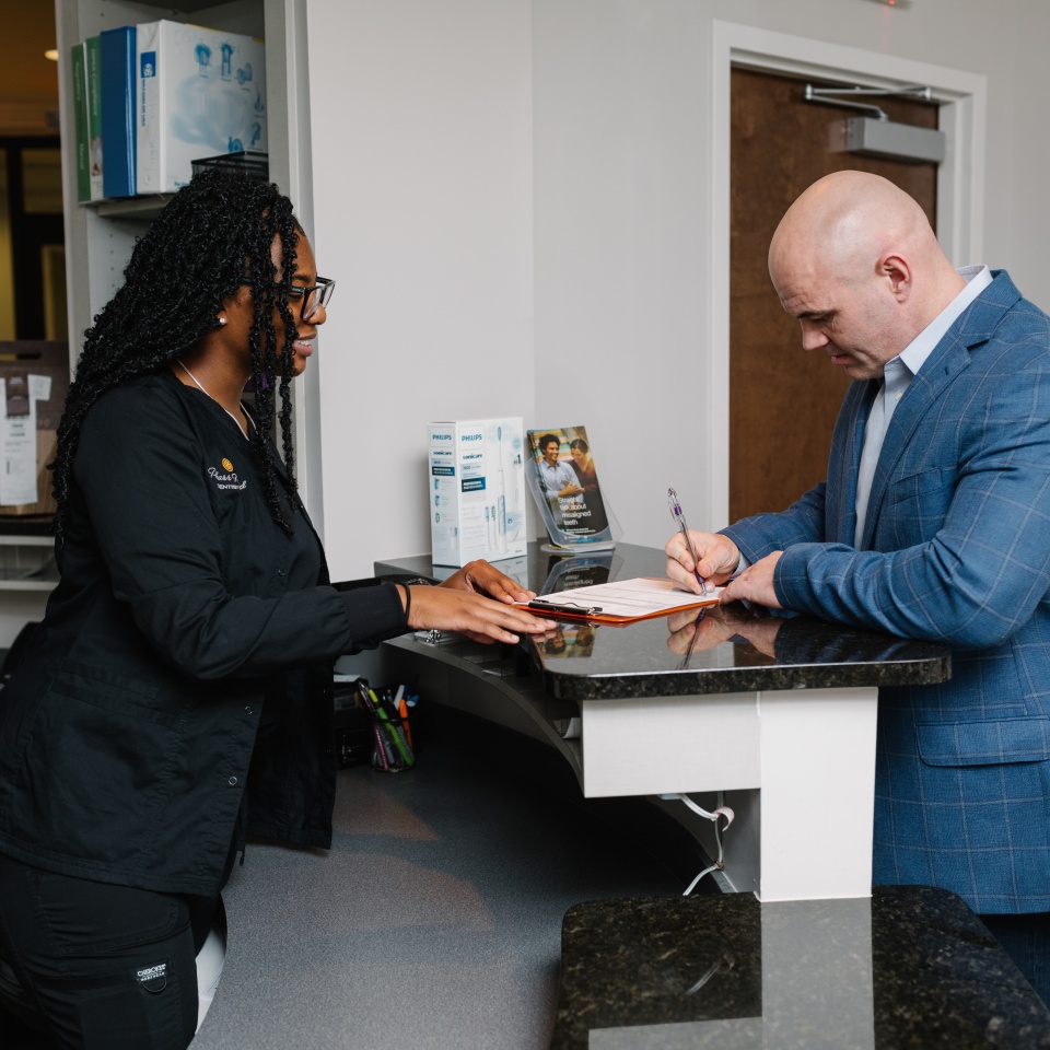 A man in a blue suit fills out paperwork at a reception desk while a staff member in black scrubs stands across from him.
