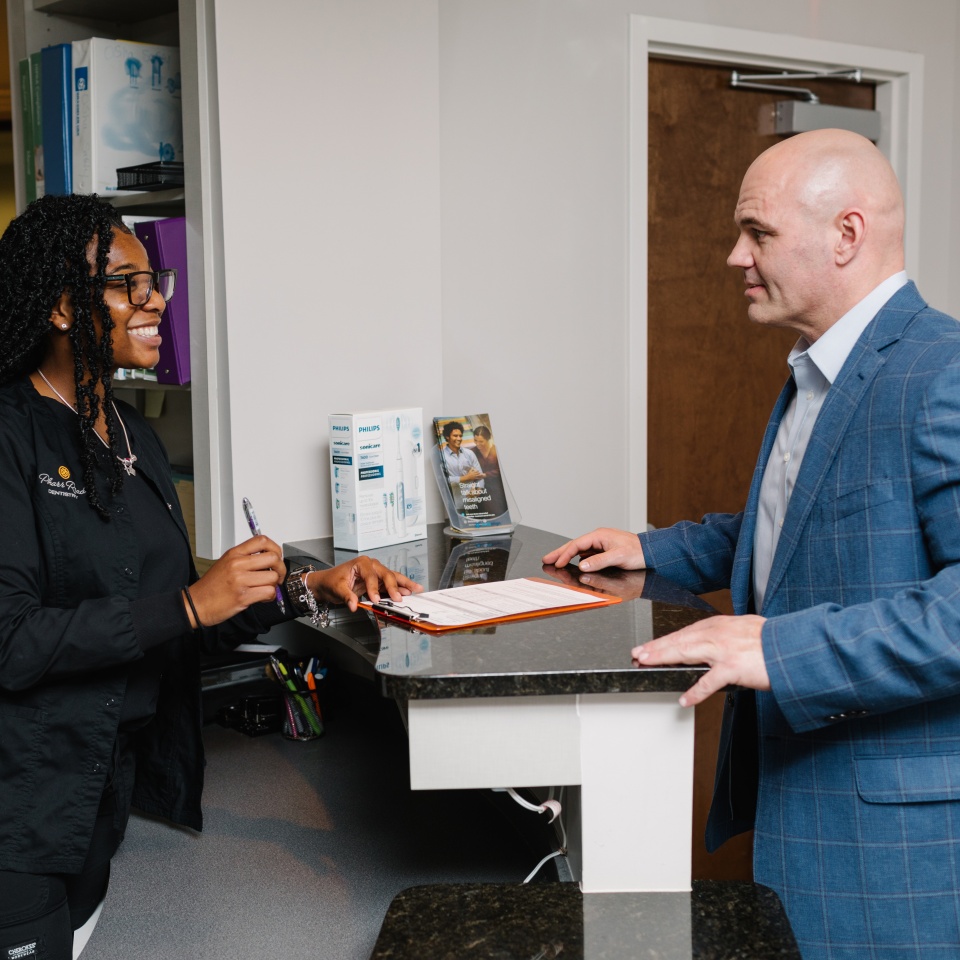 A man in a blue suit stands at a reception desk, speaking with a woman in black scrubs who is holding a pen and clipboard.