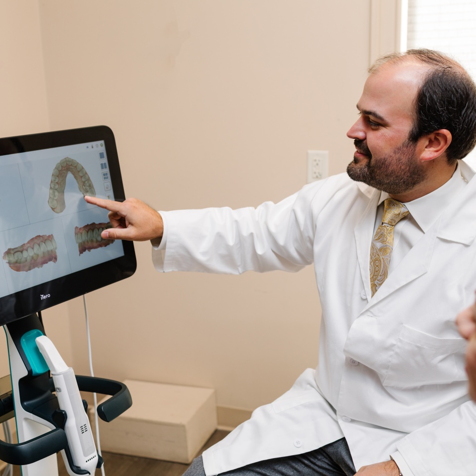A dentist in a white coat points to a 3D dental scan displayed on a monitor during a consultation in an examination room.