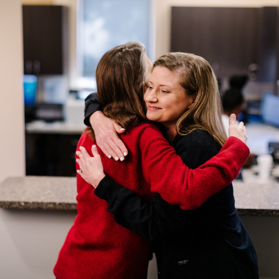 Two women hug each other in an office setting, one wearing a red jacket and the other in black, with desks and computers visible in the background.