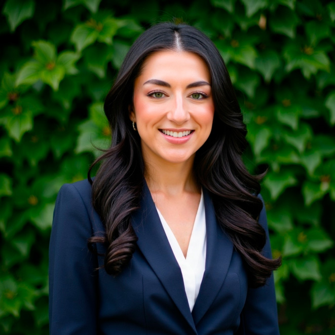 A woman with long dark hair, wearing a navy blazer and white blouse, stands outdoors in front of a leafy green background, smiling at the camera.