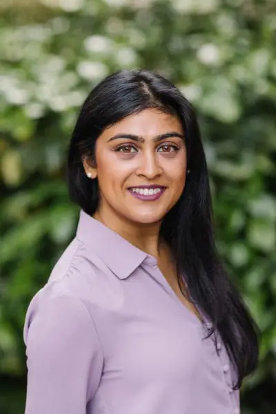 A woman with long dark hair, wearing a light purple blouse, stands in front of a blurred green leafy background and smiles at the camera.
