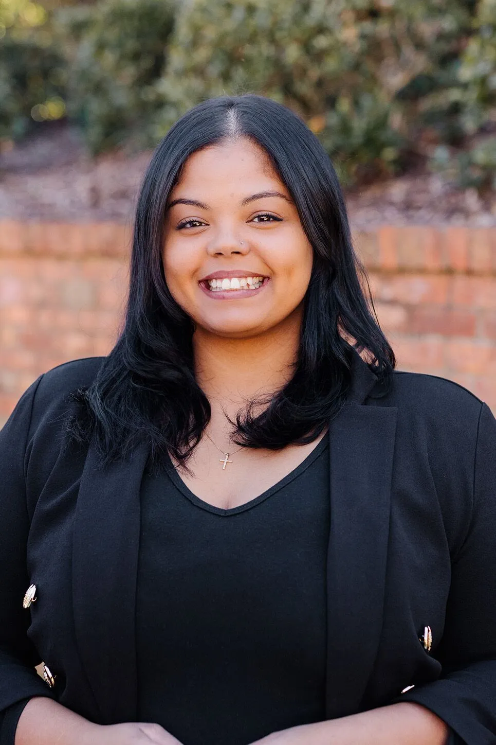 A woman with long black hair, wearing a black blazer and top, smiles at the camera outdoors with a brick wall and greenery in the background.