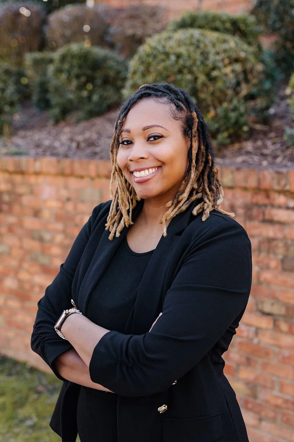 A woman with blonde-tipped dreadlocks, wearing a black blazer and top, stands outdoors in front of a brick wall, smiling with arms crossed.