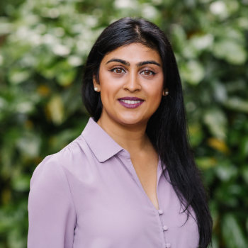 Woman with long black hair wearing a light purple blouse, standing outdoors in front of a leafy green background, looking at the camera and smiling slightly.