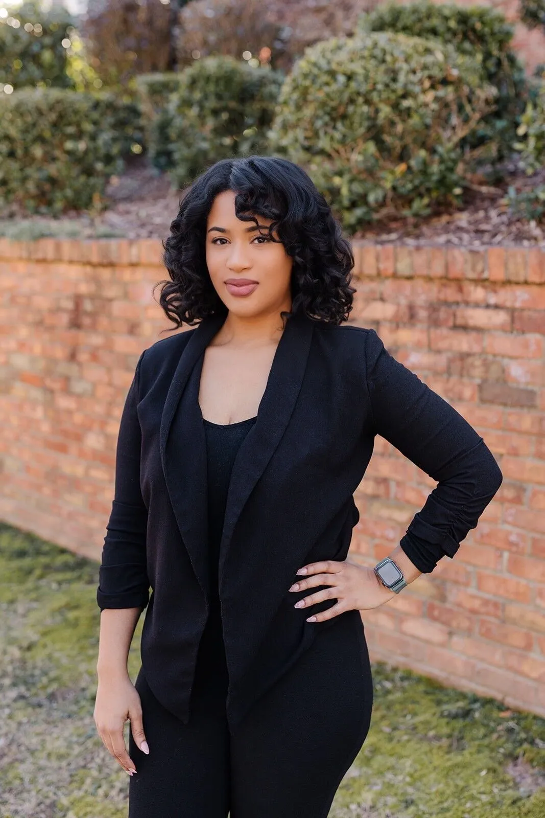 A woman with curly black hair wearing a black outfit stands outdoors in front of a brick wall and greenery, with one hand on her hip.