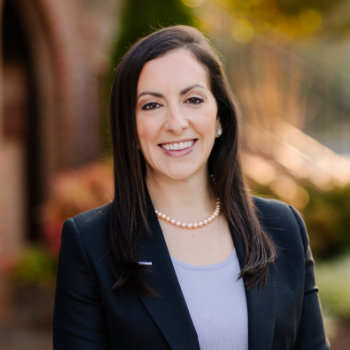 A woman with long dark hair wearing a black blazer, light top, and pearl necklace stands outside, smiling at the camera.