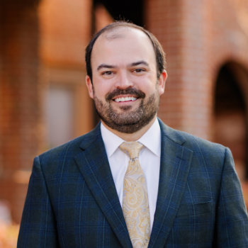 A man in a checked blazer, white shirt, and patterned tie stands outdoors in front of a brick building, smiling at the camera.