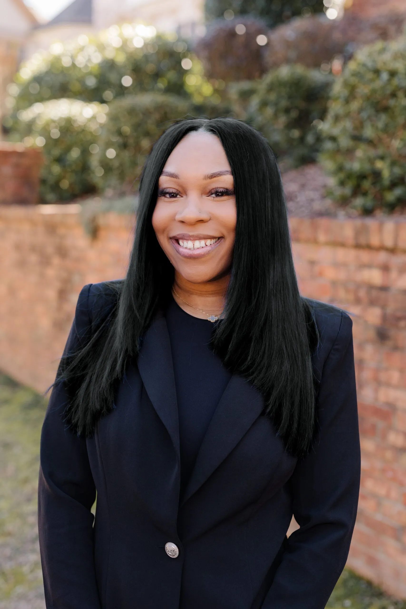 A woman with long black hair, wearing a dark blazer, stands outside in front of a brick wall and greenery, smiling at the camera.