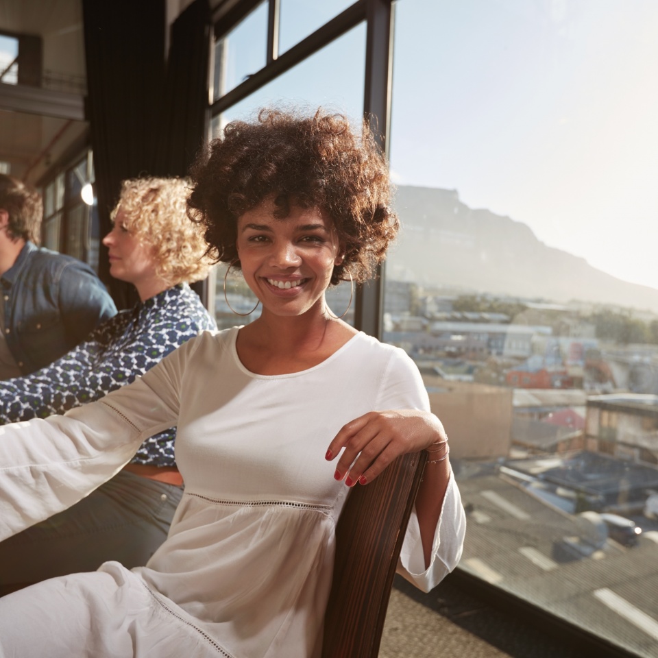 Portrait of happy young african woman sitting at a meeting with colleagues in background. Smiling creative professional looking at camera.