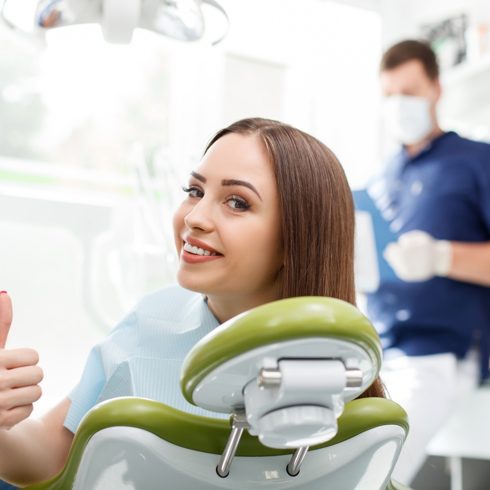 Attractive healthy woman is visiting her dentist. She is sitting in chair and looking back with joy. The lady is giving thumb up and smiling. Her dentist is standing and preparing for treating