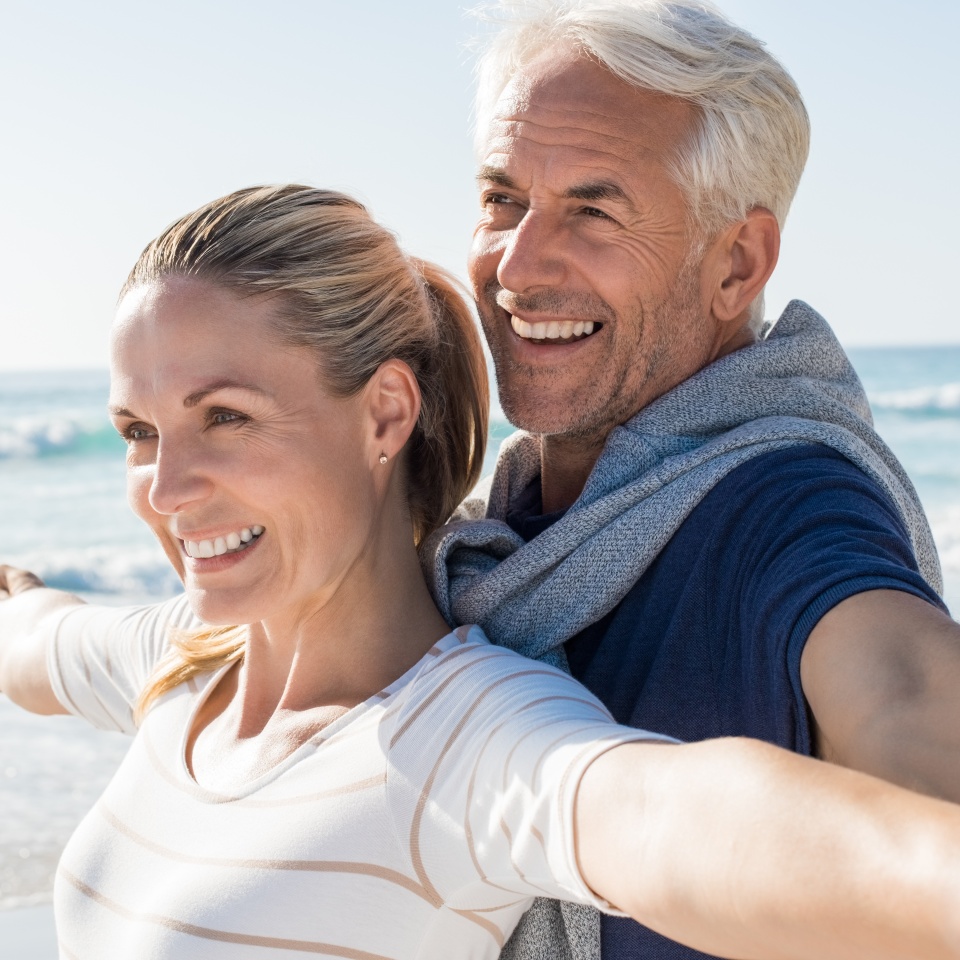 Happy senior couple standing on beach with arms outstretched and looking away. Happy couple at beach on a bright sunny day. Retired husband and smiling wife thinking about their future.