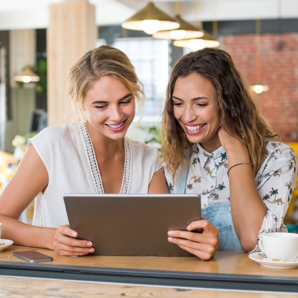 Two happy smiling young women in a cafeteria using digital tablet. Happy girls friends sitting at coffee shop looking at tablet. Happy multiethnic women drinking coffee while using tablet.