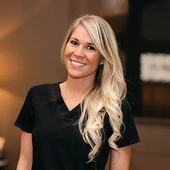 Woman with long blonde hair wearing a black scrub top, standing indoors and smiling at the camera.
