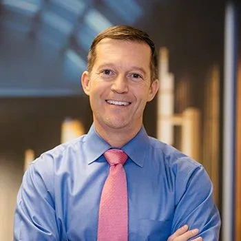 Man in a blue dress shirt and pink tie stands with arms crossed, smiling, in an office setting with blurred background.