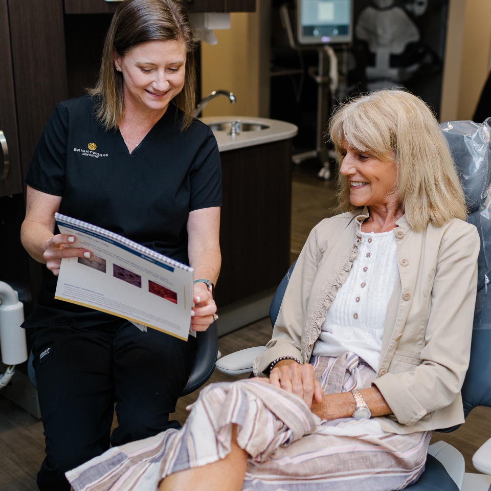A dental professional shows a brochure to a smiling older woman seated in a dental chair in a clinic setting.
