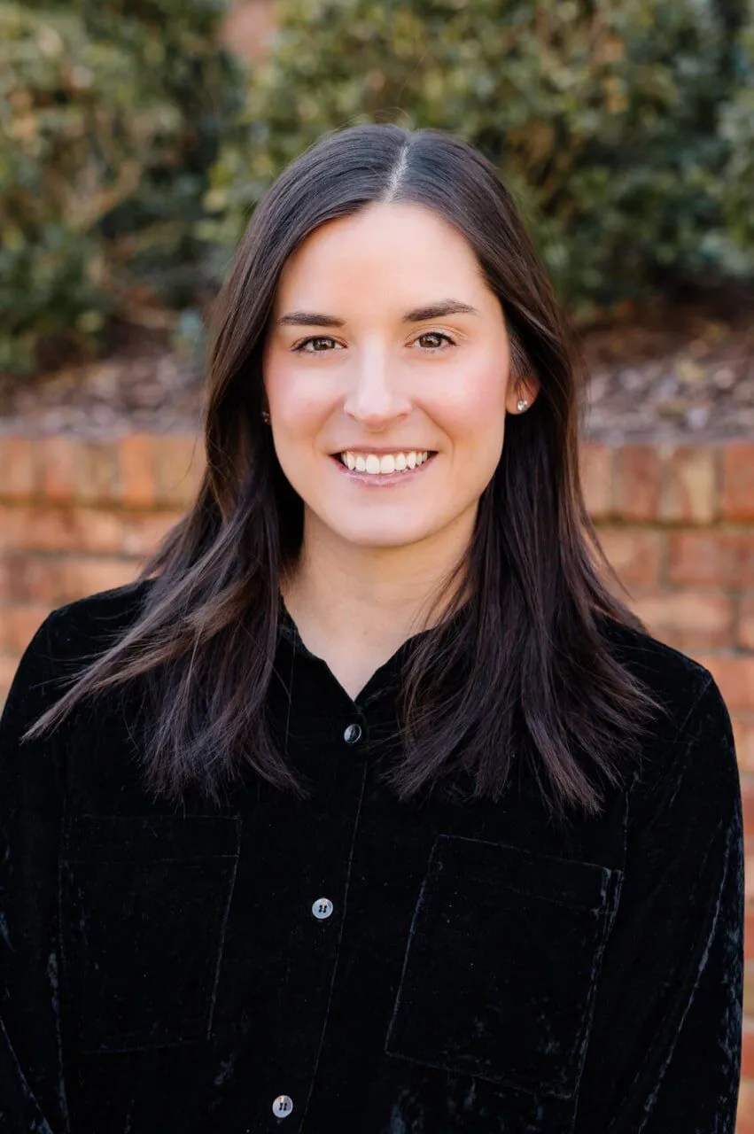 A woman with straight dark brown hair wearing a black top stands outdoors in front of a brick wall and greenery, smiling at the camera.