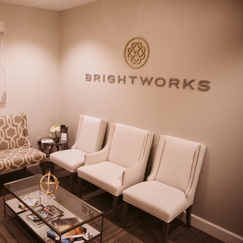 A waiting room with three beige chairs, a patterned armchair, a glass coffee table with magazines, and a "BRIGHTWORKS" sign on the wall.