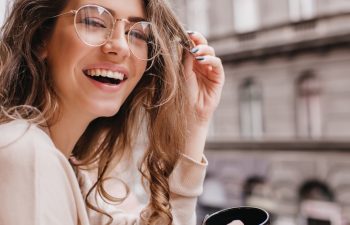 Close-up portrait of laughing brunette girl in beige sweater drinking coffee on city background. Lovable lady standing near window and looking at beautiful building with cup of tea.