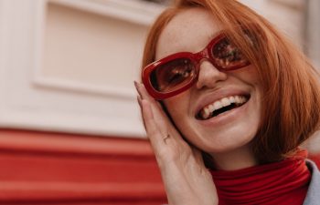 Close-up portrait of young beautiful lady posing outdoors. Pretty red-haired girl wearing turtleneck and blue shirt, adjusting trendy sunglasses and smiling against bright wall background