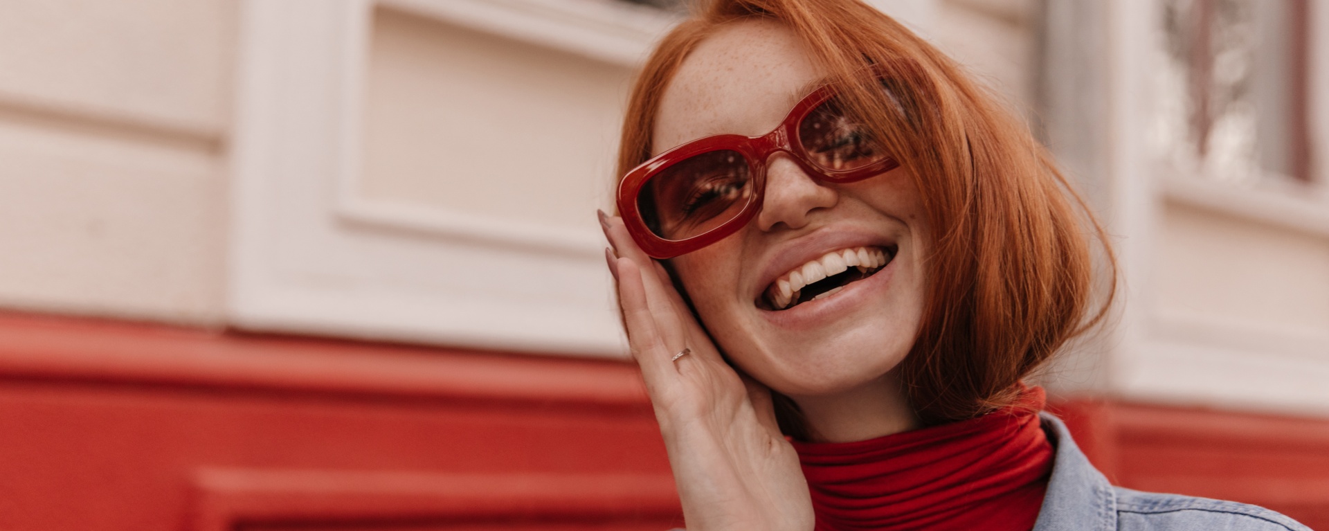 Close-up portrait of young beautiful lady posing outdoors. Pretty red-haired girl wearing turtleneck and blue shirt, adjusting trendy sunglasses and smiling against bright wall background