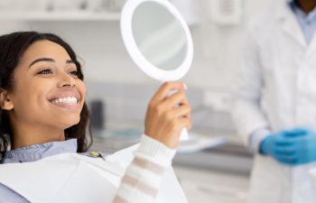 Happy Black Female Patient Looking At Mirror After Dental Treatment In Clinic, Cheerful African American Woman Sitting In Chair In Stomatological Cabinet And Enjoying Her Beautiful Smile, Closeup