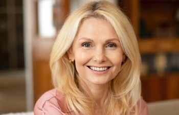 Portrait Of Blonde European Mature Woman Posing Smiling To Camera Sitting On Sofa At Home. Lady Resting On Cozy Couch In Modern Living Room Indoor. Ageless Beauty And Elegance Concept