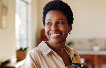 Happy Brazilian woman looking outside the window in her home kitchen, smiling and holding an enamel mug of plain coffee. Mature, afro-haired woman standing next to a breakfast table.