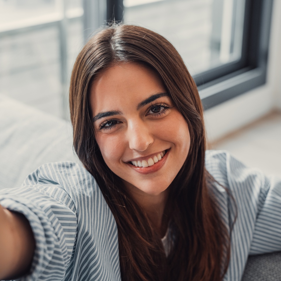 Happy attractive young adult woman housewife sitting on sofa looking at camera in modern cozy home, smiling single 20s lady beautiful face taking a selfie alone enjoy wellbeing posing for portrait