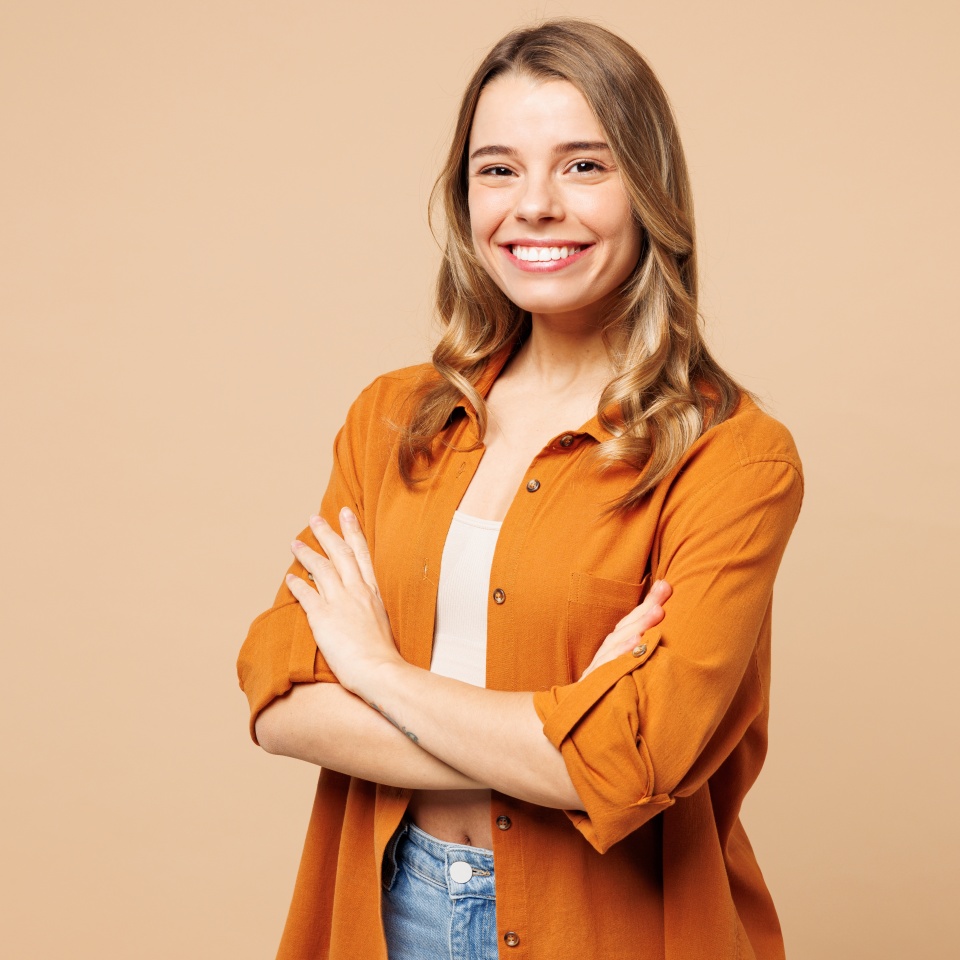 Side view young smiling Caucasian woman she wear orange shirt casual clothes hold hands crossed folded look camera isolated on plain pastel light beige background studio portrait. Lifestyle concept