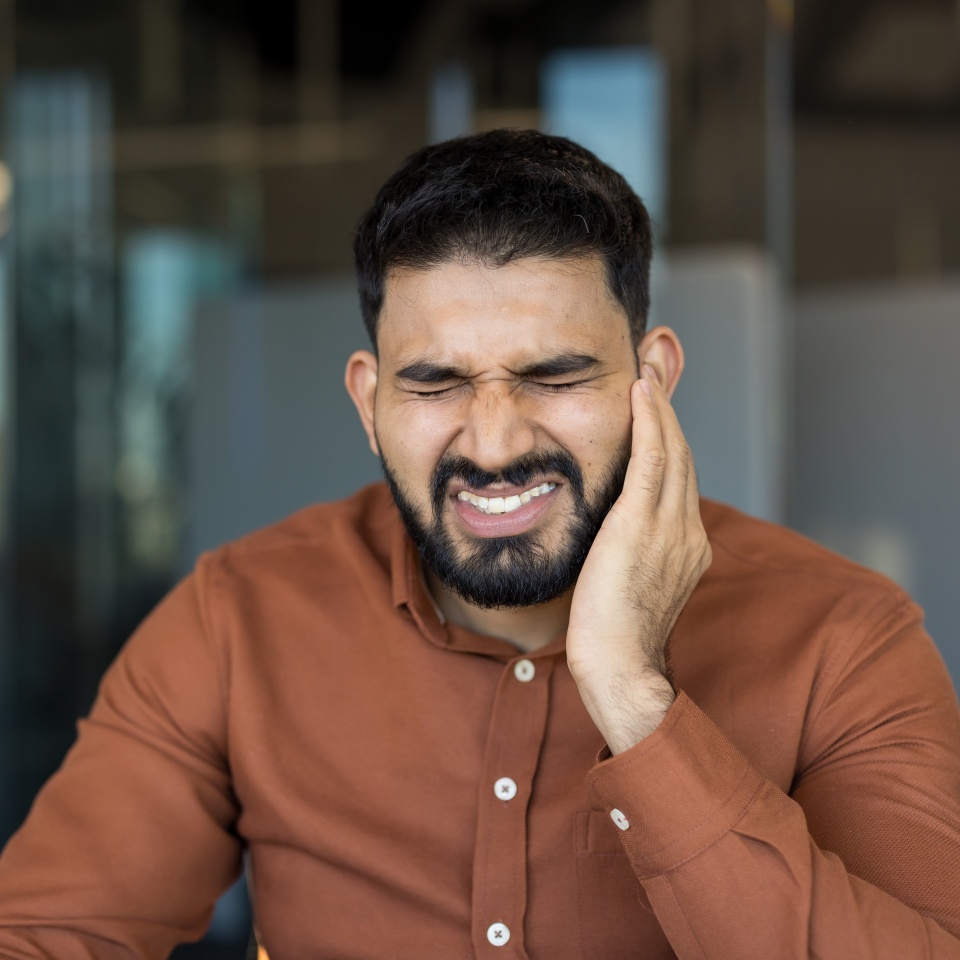 Young middle eastern indian man grimacing in pain, clutching his cheek from a severe toothache or jaw inflammation, urgent dental problem needing immediate care