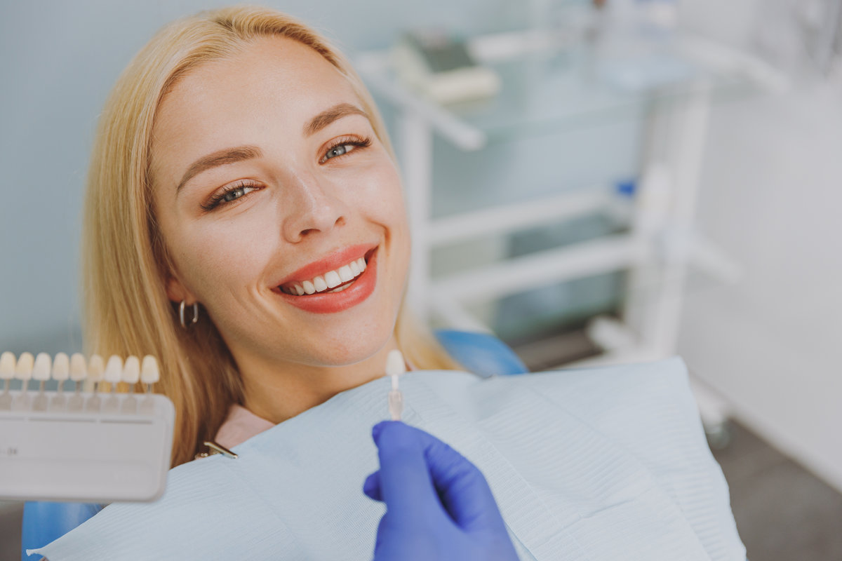 Close up young smiling woman choosing veneers enamel color palette with stomatologist compare teeth shade sit at dentist office chair indoor light modern cabinet Healthcare enamel whitening treatment
