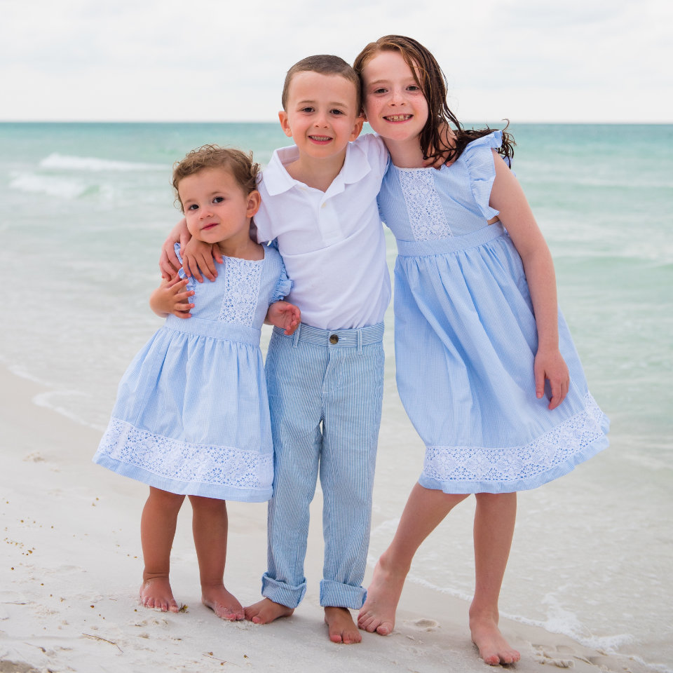 Three young children stand together on a beach. The boy in the middle wears a white shirt and light blue pants. The two girls wear matching blue dresses. The ocean is in the background.