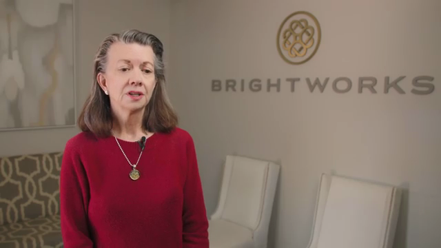 A woman in a red sweater stands in front of a wall with the "BRIGHTWORKS" logo and three white chairs.
