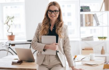 Confident business expert. Attractive young smiling woman in smart casual wear holding digital tablet and looking at camera while leaning on desk in creative office